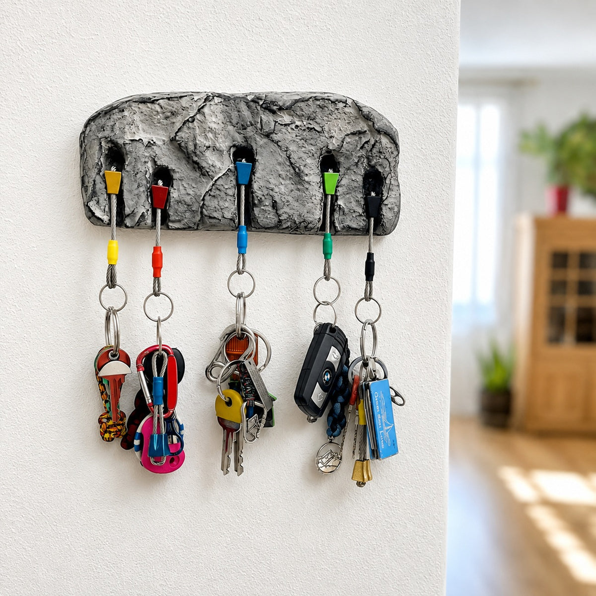 A gray faux-stone key rack mounted on a white wall, holding five colorful keychains and several keys, with a blurred home interior in the background.