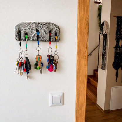 A textured gray stone-look key rack mounted on a white wall, holding several colorful keychains and keys in a home entryway.