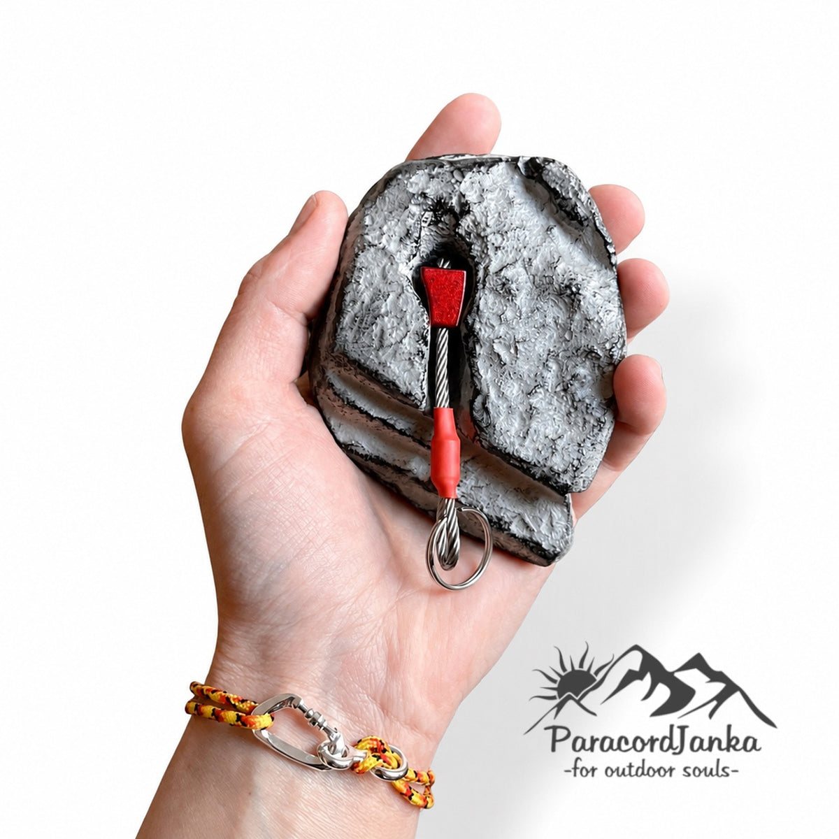 A hand holds a textured gray rock-like object with a small red and silver cord anchor tool inserted into it. The wrist also wears a yellow and orange braided cord bracelet.