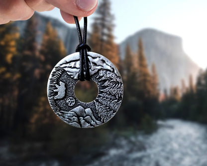 A hand holding up a Hiking Mountain Necklace Gift with a mountain peaks background.