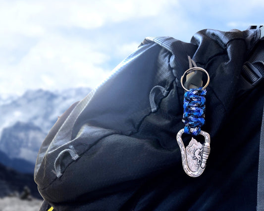 May include: A blue and white paracord keychain with a silver carabiner clip. The clip features a mountain climber design. The keychain is attached to a black backpack, with a mountain range visible in the background.