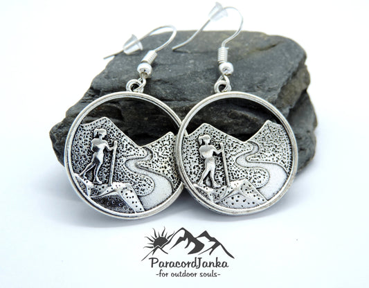 May include: Silver earrings with a hiker silhouette on a mountain path design. The earrings are circular and have a hook closure.