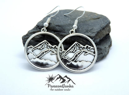 May include: Silver-toned mountain range earrings. Each earring features a circular pendant with a detailed mountain scene. The earrings have a hook closure. The ParacordJanka logo is visible.