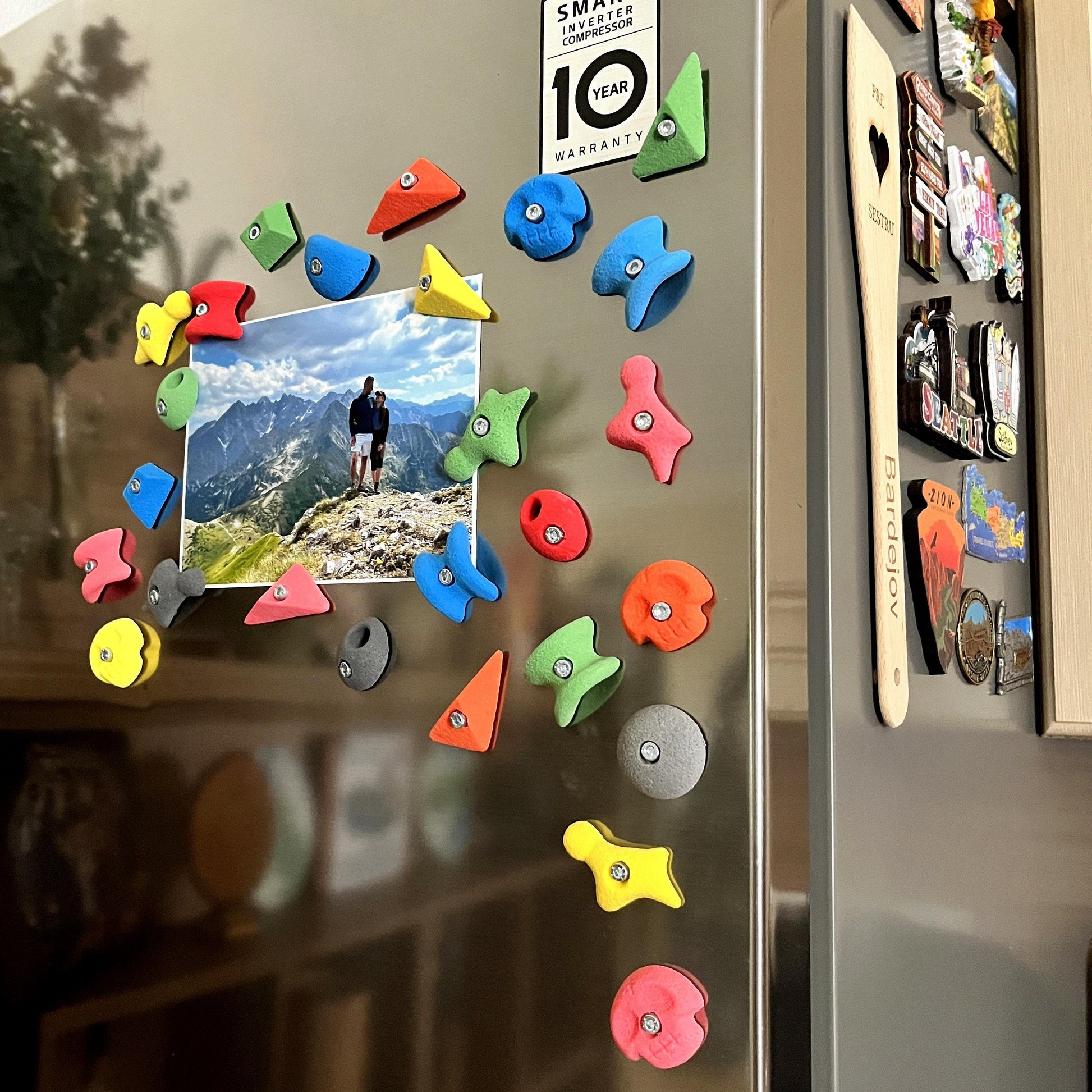 May include: A collection of colorful climbing hold magnets in various shapes and sizes, attached to a refrigerator door. The magnets are in shades of red, blue, green, yellow, and orange. A photograph is also attached to the door.