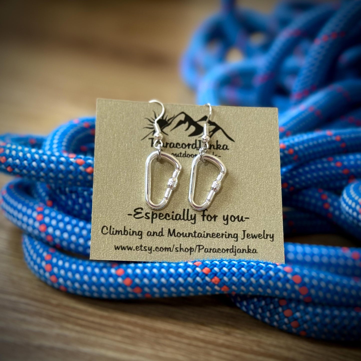 May include: Silver carabiner-shaped earrings on a tan card with the text "-Especially for you- Climbing and Mountaineering Jewelry". The earrings are displayed against a backdrop of blue climbing rope.