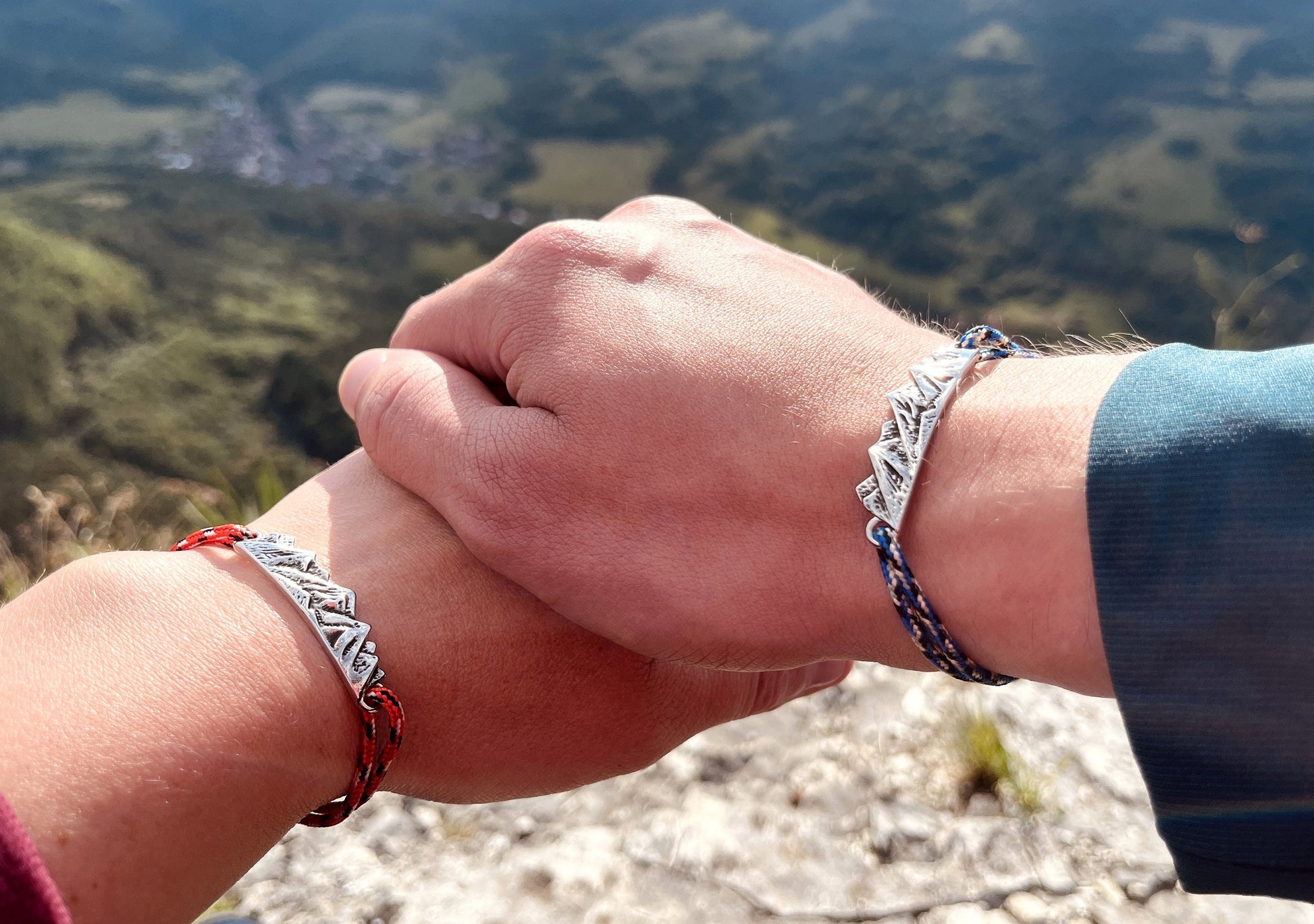 May include: Two people wearing matching bracelets with a mountain range design. The bracelets are made of red and blue cord and have a silver charm. The bracelets are on the wrists of the people's hands, which are clasped together.