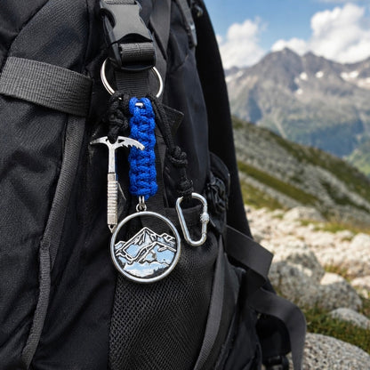 A close-up of a black hiking backpack with a blue paracord keychain and silver mountain emblem hanging from a strap, set against a blurred mountain landscape.