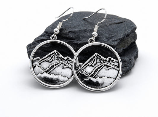 A pair of silver-toned round drop earrings with an engraved mountain landscape design, displayed on a dark slate rock against a white background.