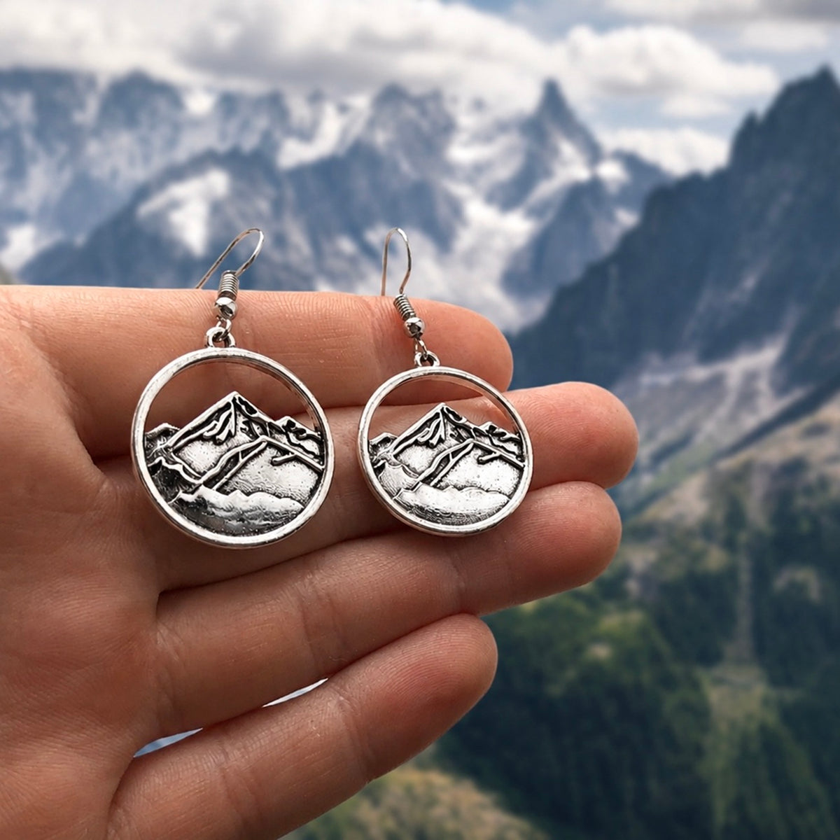 A pair of silver-toned round drop earrings with an engraved mountain landscape design, held in a person's hand against a blurred mountain backdrop.