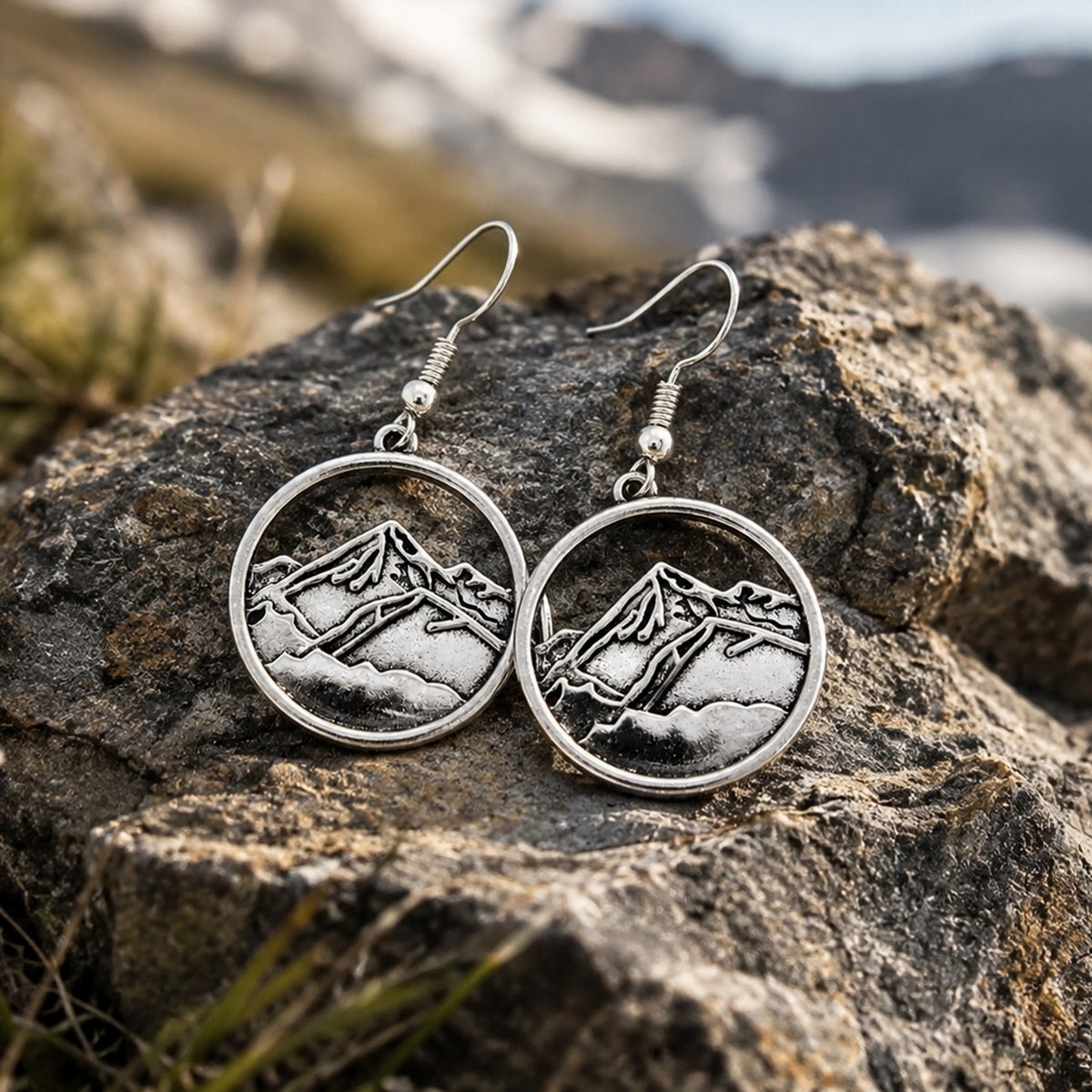 A pair of silver-toned round drop earrings with an engraved mountain landscape design, resting on a large textured rock with a blurred mountain background.
