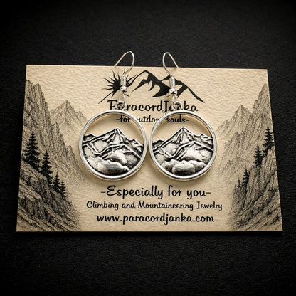 A pair of silver-toned round hoop earrings featuring an embossed mountain landscape design, displayed on a branded beige card against a dark textured background.