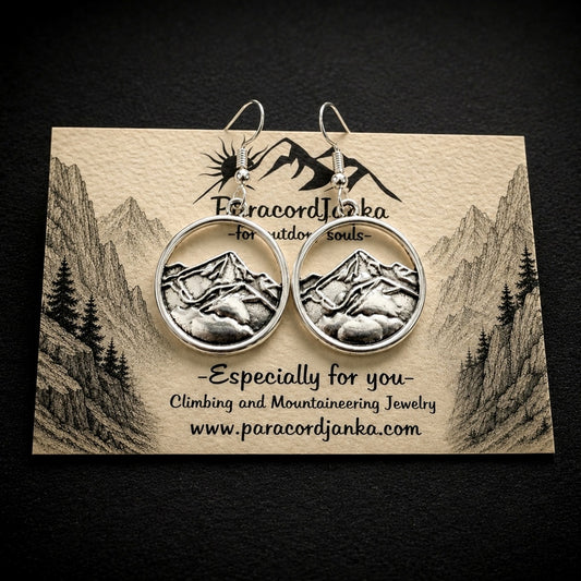 A pair of silver-toned round hoop earrings featuring an embossed mountain landscape design, displayed on a branded beige card against a dark textured background.
