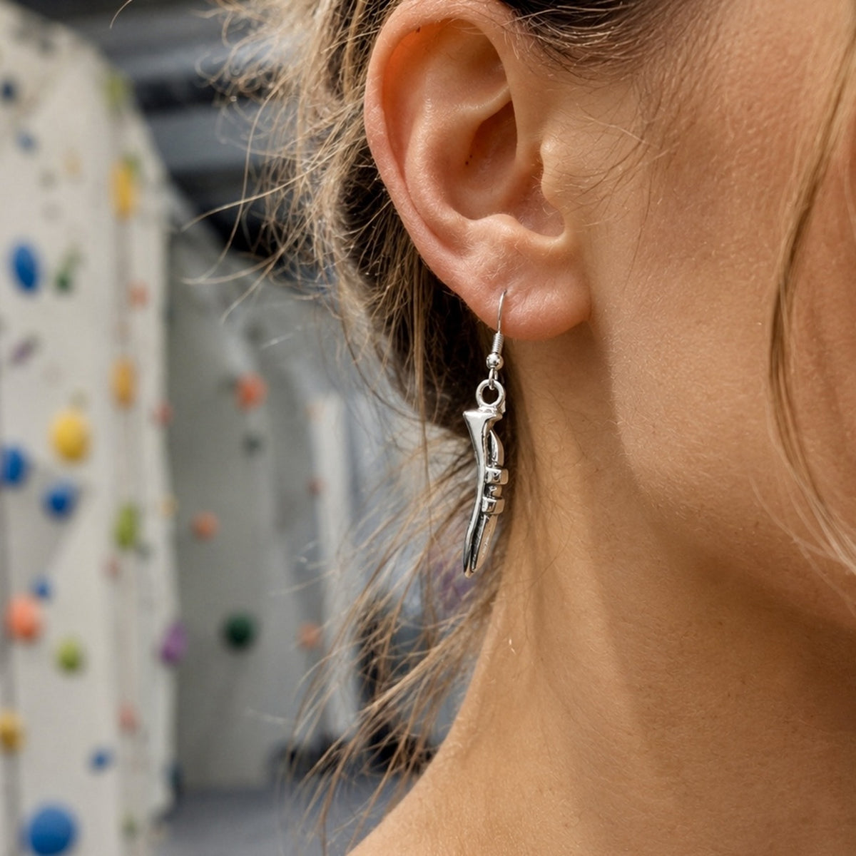 A close-up of a woman's ear wearing a silver dangling earring shaped like a stylized bone or claw, with a blurred indoor climbing wall in the background.