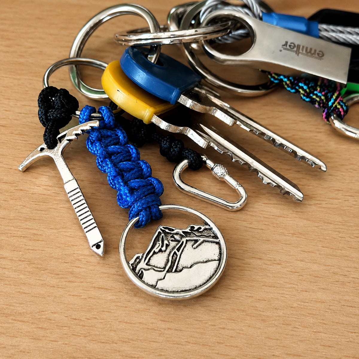 A close-up of a keychain set on a light wood surface, featuring silver metal keys and rings, a blue paracord-wrapped pick, a silver mountain medallion, a black and blue paracord charm, and a yellow keyring accent.