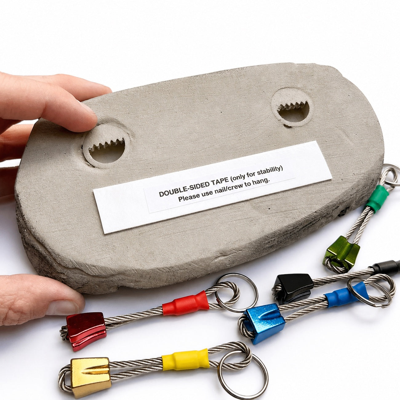 A gray concrete anchor block with a white label reading 'DOUBLE-SIDED TAPE (only for stability) Please use nailcrew to hang' is held by two hands, with several colorful metal cable anchors and key rings displayed in front on a white background.