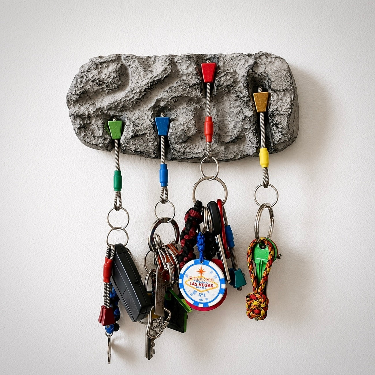 A textured gray rock-shaped key holder mounted on a white wall, featuring five colorful hook inserts in green, blue, red, yellow, and brown, holding various keys and keychains.