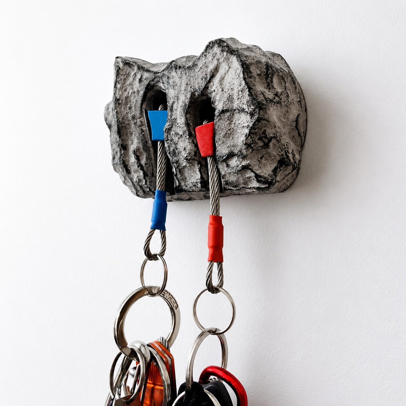 A novelty rock-shaped wall hook holder mounted on a white wall, with two metal cable keychains hanging into its carved holes, one with a blue cap and one with a red cap.