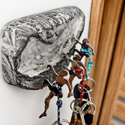 A close-up of a black and white rock wall hook holding several metal keychains with colorful rope accents, against a white wall with a wooden door frame visible on the right.