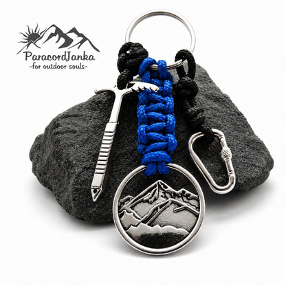 A mountain climbing keychain with a blue and black paracord wrap, featuring a silver-tone mountain medallion, ice axe, and carabiner, displayed on a dark gray rock with the ParacordJanka logo in the top left corner.