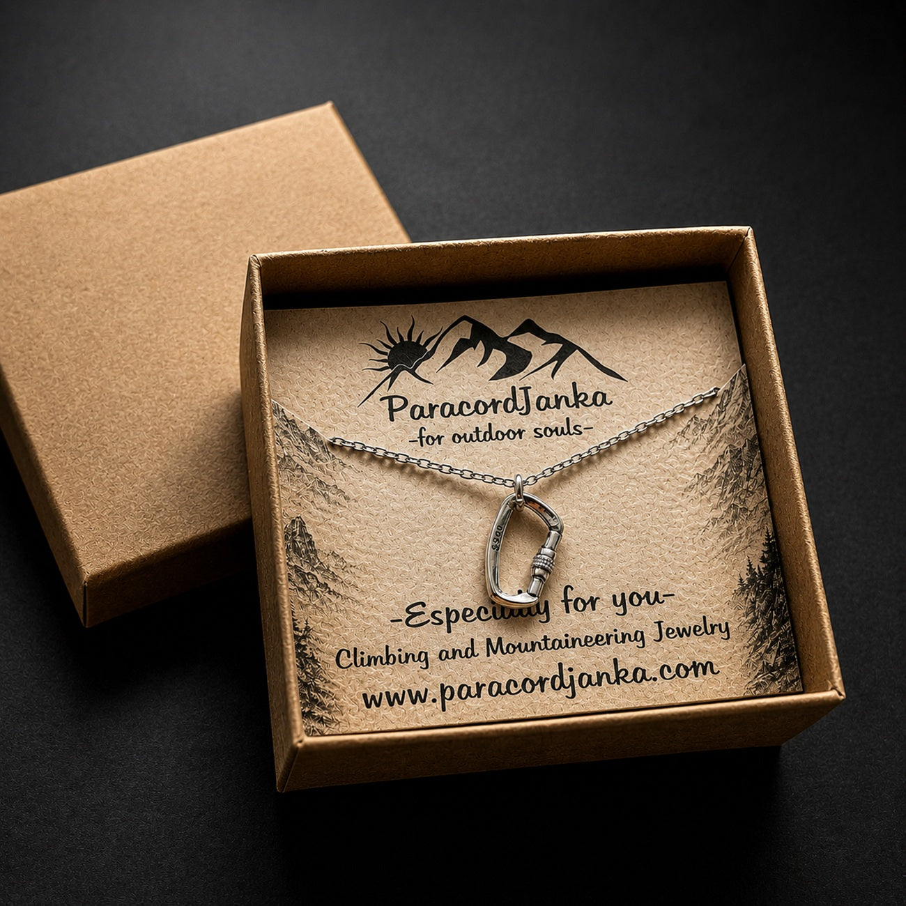 A silver mountain-shaped pendant necklace displayed on a kraft paper card inside an open brown gift box. The card is branded ParacordJanka with mountain graphics and text about outdoor/climbing jewelry.