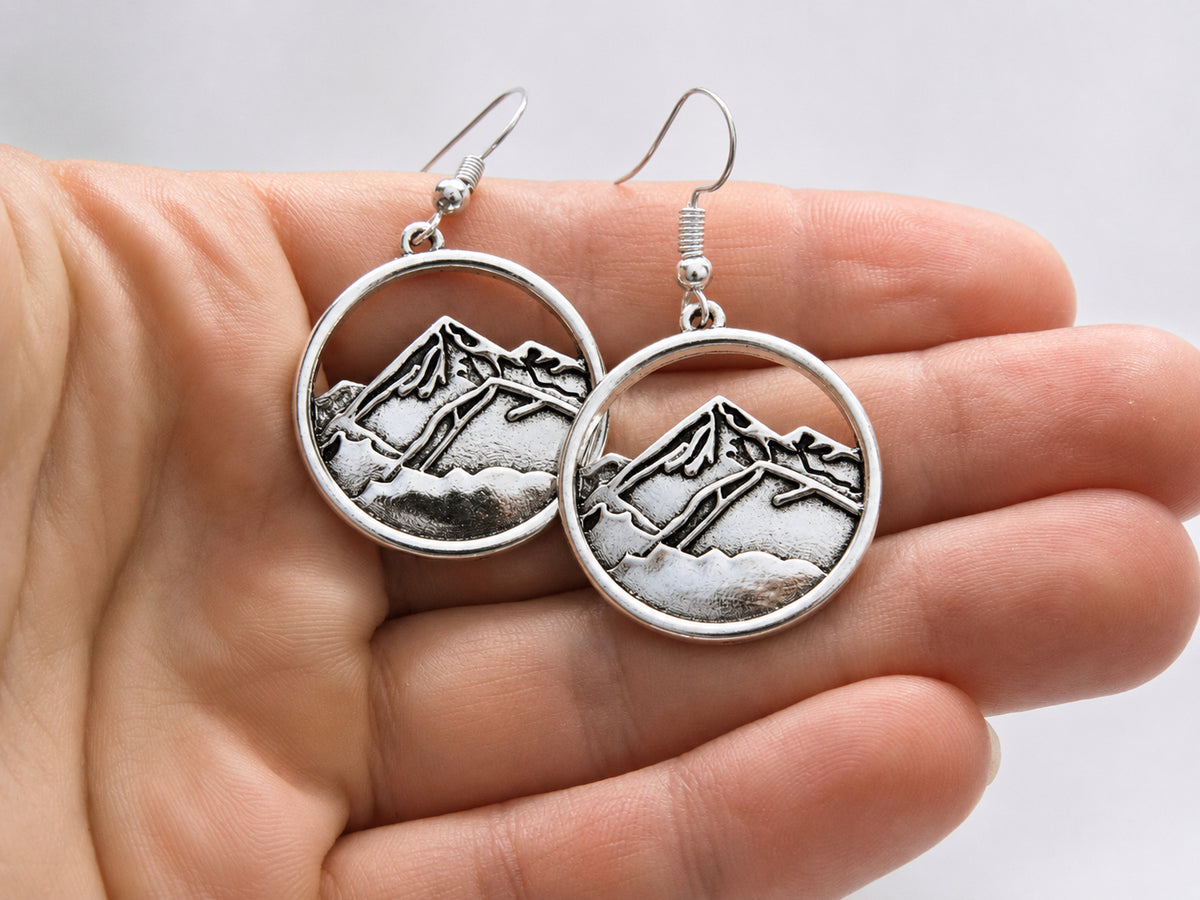 A pair of silver-tone round drop earrings with an engraved mountain landscape design, held in a person's hand against a light gray background.
