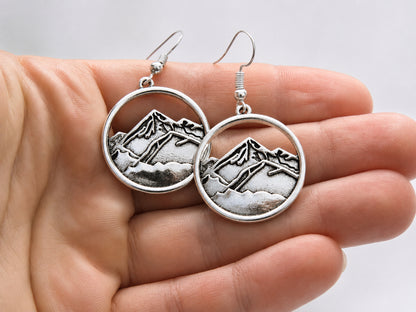 A pair of silver-tone round drop earrings with an engraved mountain landscape design, held in a person's hand against a light gray background.