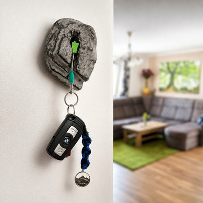 A novelty keychain holder shaped like a gray rock mount on a white wall, holding a black car key fob and blue beaded keychain. The right side shows a blurred living room interior.