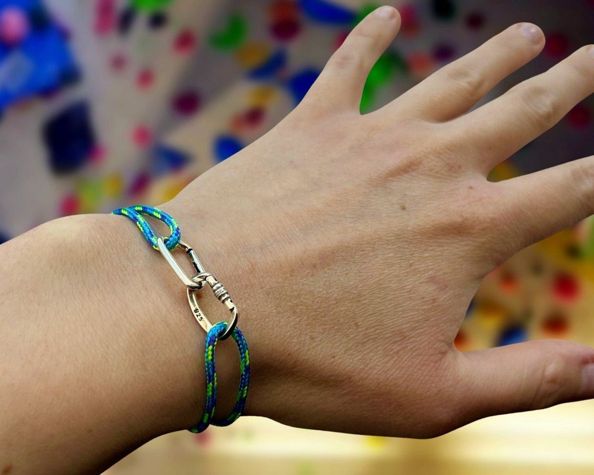 A close-up of a person's hand wearing a braided rope climbing bracelet with a silver carabiner clasp. The rope is primarily blue and green, and the clasp is silver.