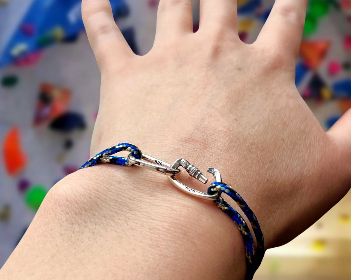 A close-up of a person's wrist wearing a silver-toned climbing bracelet with blue and white patterned woven cords, set against a blurred background of colorful climbing holds.